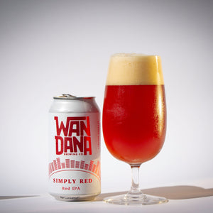 A can of Wanda Brewing Co. Simply Red Red IPA beer stands next to a tulip-shaped glass filled with amber beer and a thick head of foam, photographed on a light gray studio background.