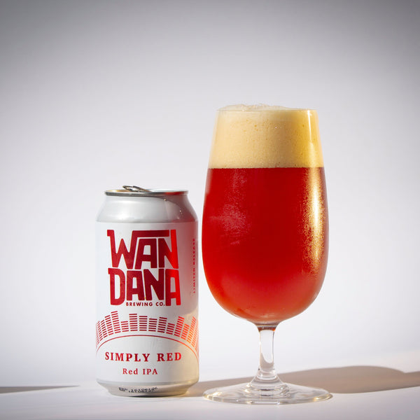 A can of Wanda Brewing Co. Simply Red Red IPA beer stands next to a tulip-shaped glass filled with amber beer and a thick head of foam, photographed on a light gray studio background.